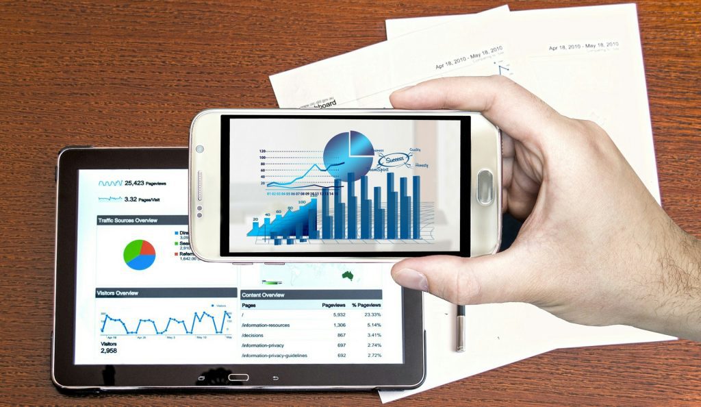 right hand holding a smart phone with analtycis displayed, and an iPad behind it on a table also showing analtycal displays