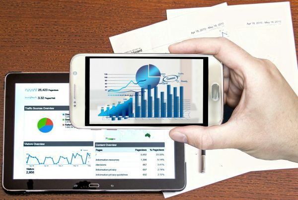 right hand holding a smart phone with analtycis displayed, and an iPad behind it on a table also showing analtycal displays